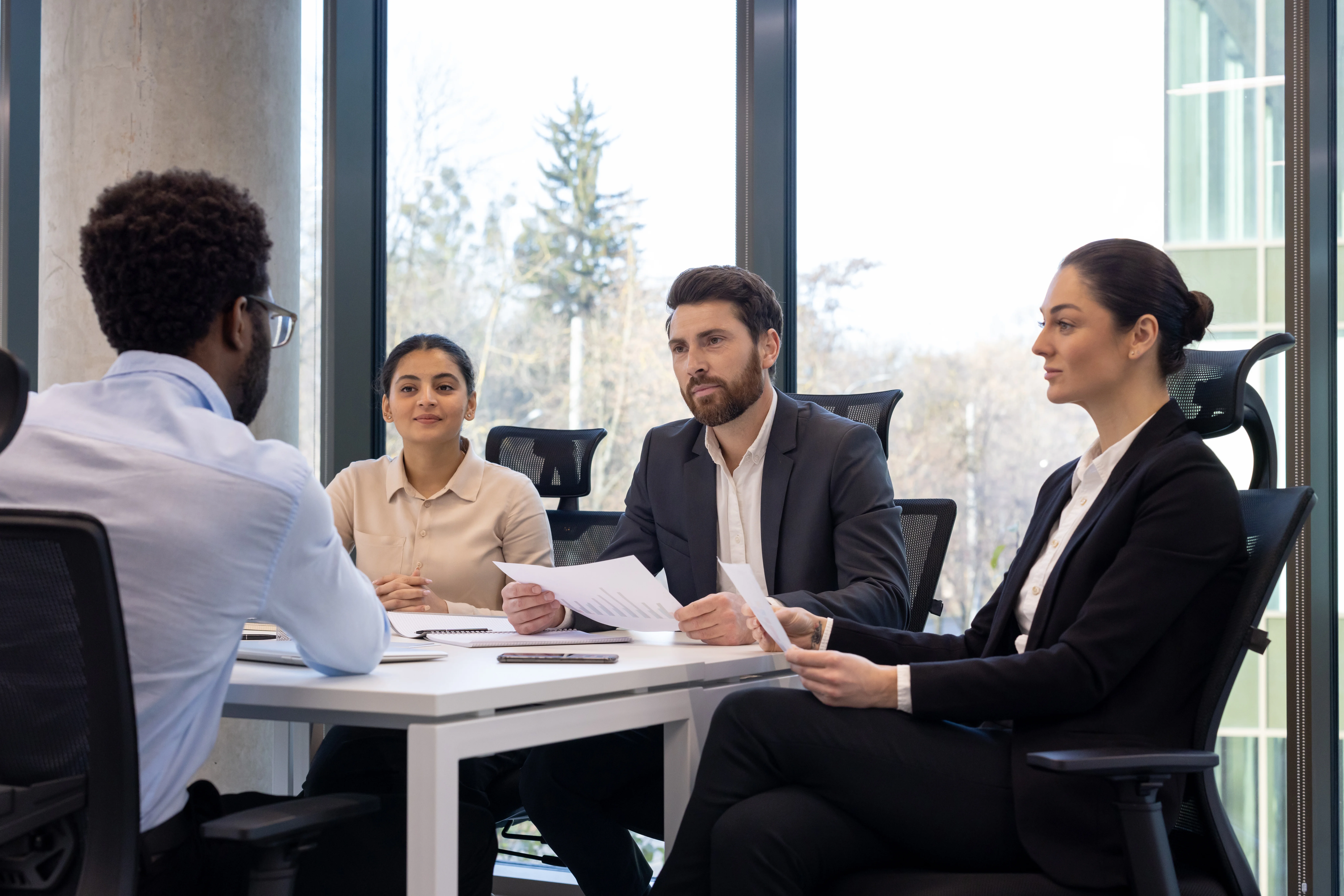 Job interview scene with diverse business team and candidate engaging professionally in modern office setting. Panel holding documents, tables equipped for corporate discussions.