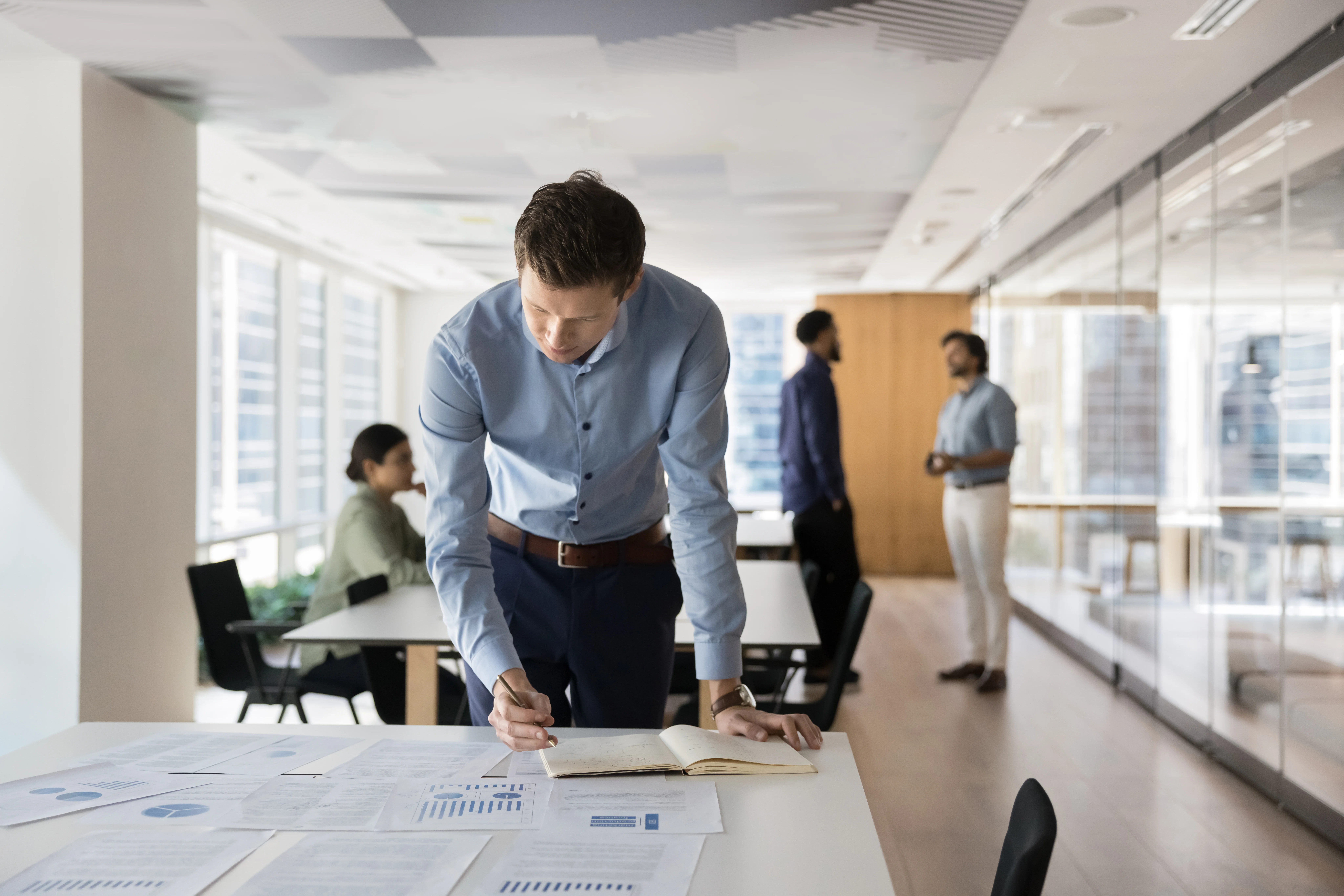 Focused busy young company owner man assessing business risk, analyzing marketing research data, statistic sales reports, studying heap of paper documents with diagrams, writing notes