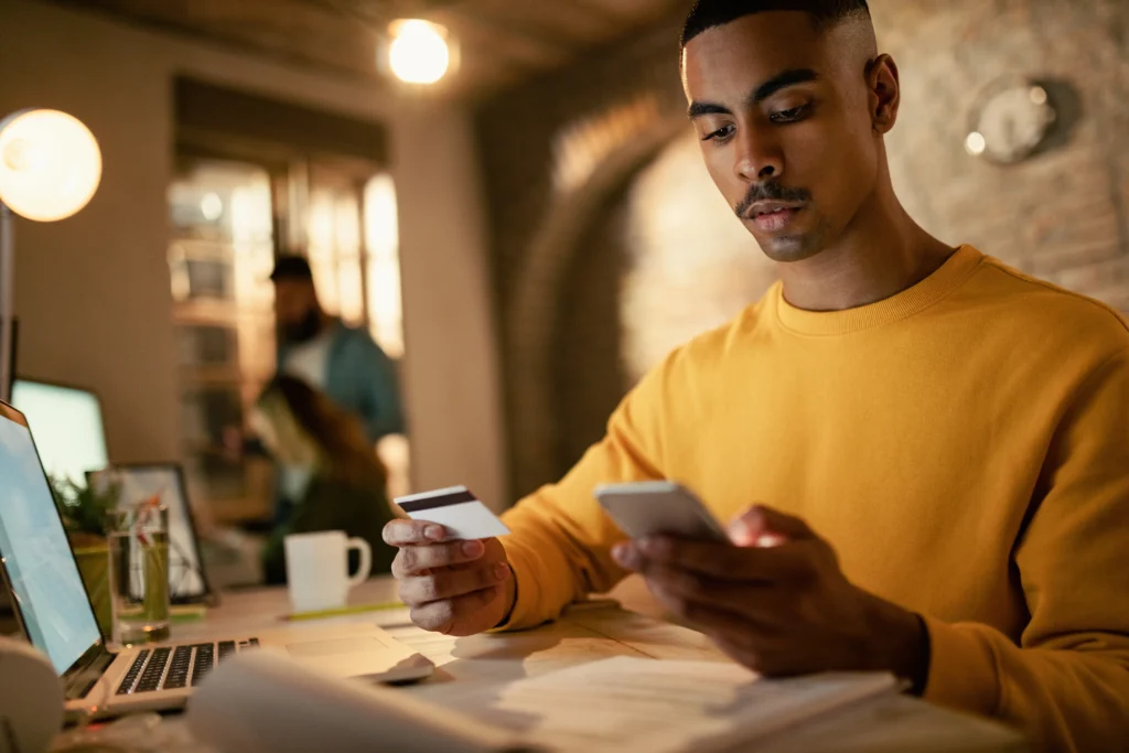 Young black businessman using credit card and smart phone while shopping online in the office - Drazen Zigic